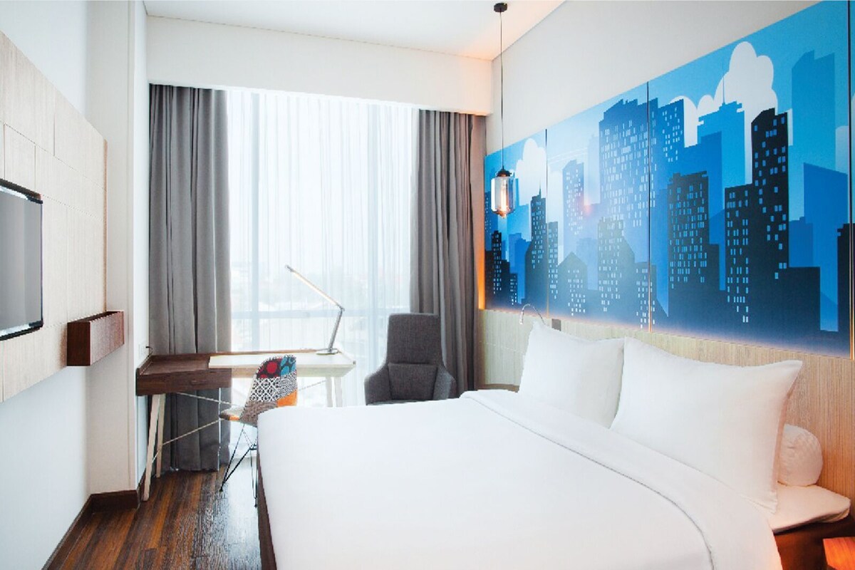 A hotel room with a large bed, a desk, a chair, a TV, and a cityscape mural on the wall.