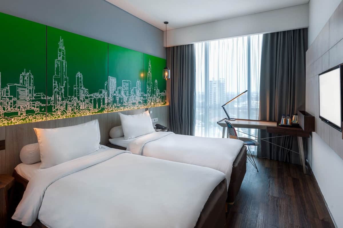 A hotel room with two beds, a cityscape mural, a desk with a chair, a large window with curtains, and a projector screen.
