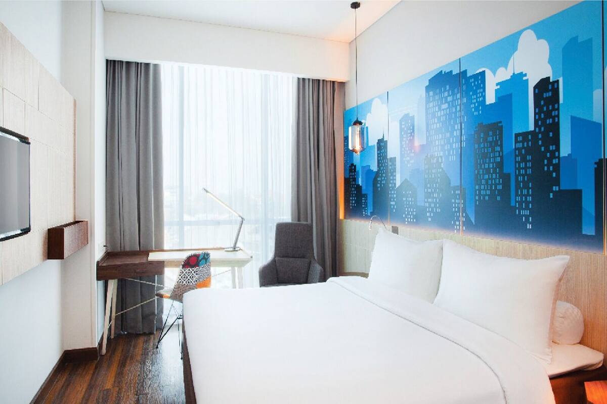 A hotel room with a large bed, a desk with a chair, a cityscape mural, and a window with curtains.