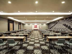 Une salle de conférence dotée de plusieurs tables et chaises, d’un sol recouvert de moquette et d’une scène avec un écran.