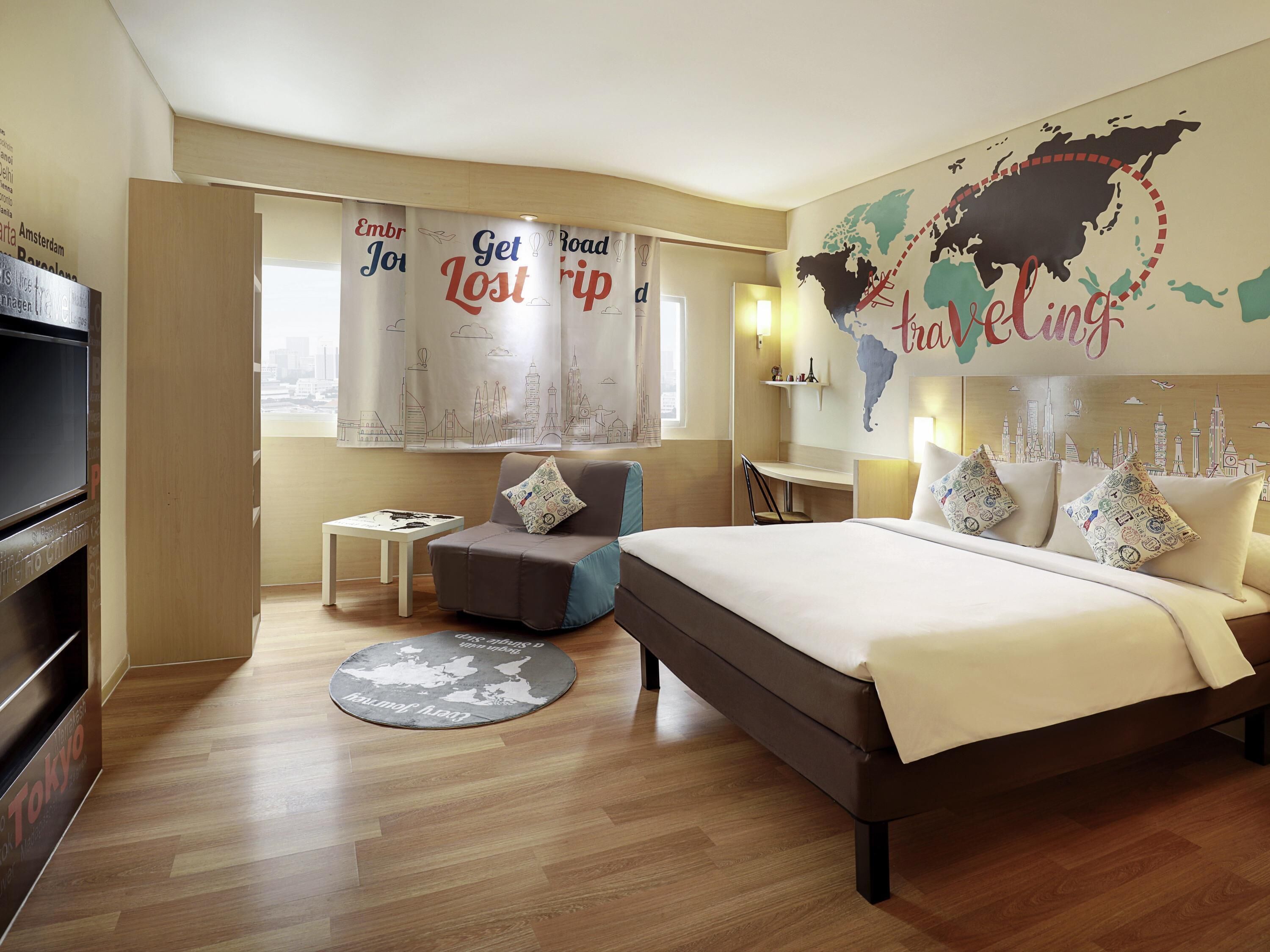 A hotel room with a bed, a desk, a chair, a TV, and a world map on the wall.
