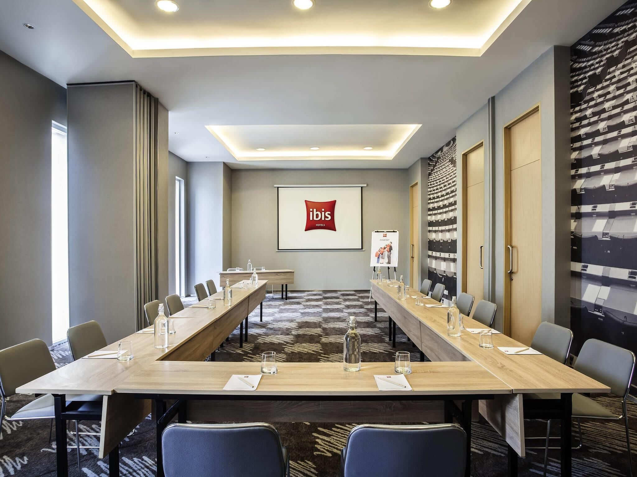 A conference room with a long table, chairs, a projector screen, and a 'ibis Hotels' sign.