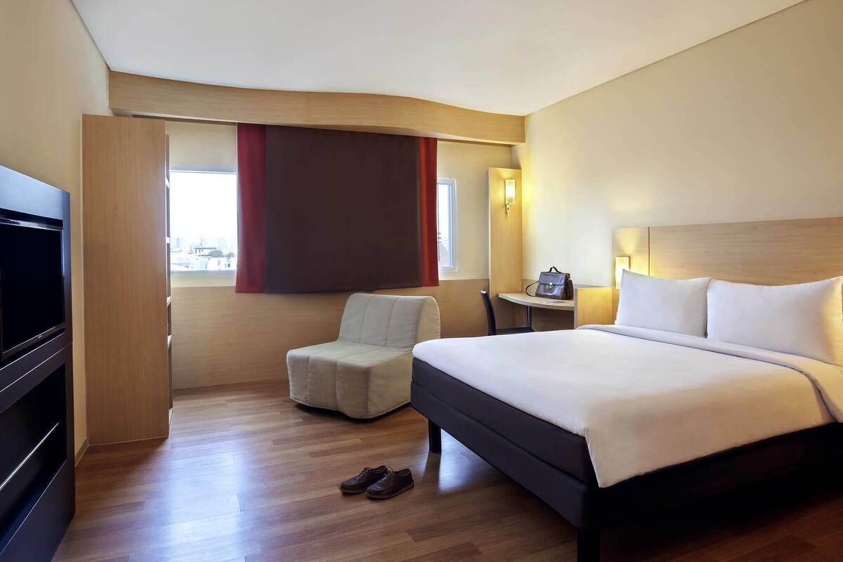 Une chambre d’hôtel avec un lit, une télévision, un bureau, une chaise et une paire de chaussures posées sur le sol.