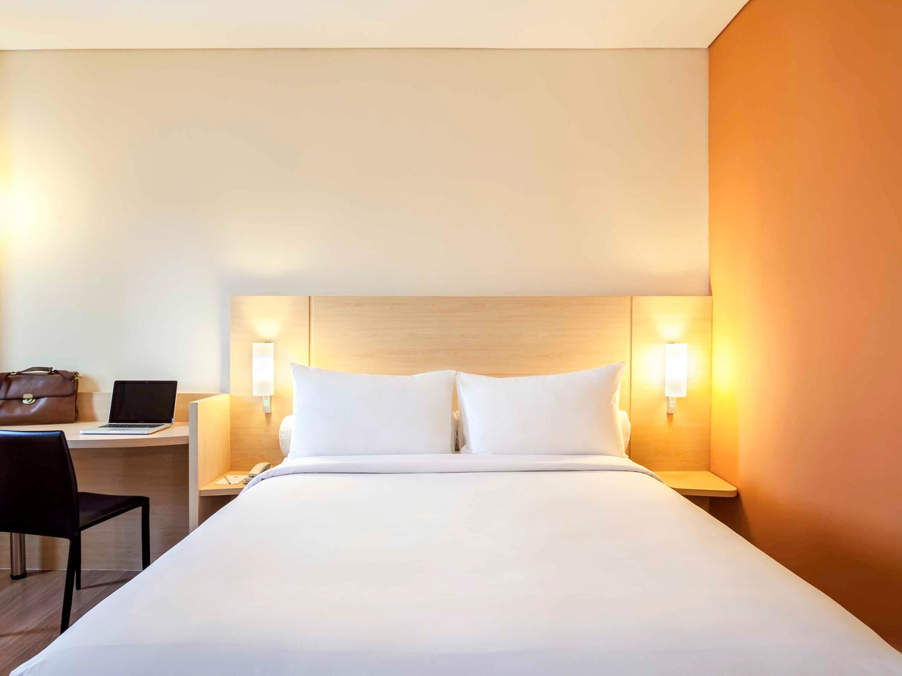 Ein Hotelzimmer mit einem großen Bett, einem Schreibtisch mit einem Laptop, einem Stuhl und einer Handtasche.