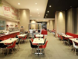 A modern restaurant interior with multiple dining tables and red chairs, a food rack, and a counter with various dishes.