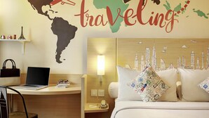 Ein Hotelzimmer mit Wandbild einer Weltkarte, einem Laptop auf dem Schreibtisch und einem Bett mit Kopfkissen im Stadtbild-Design.