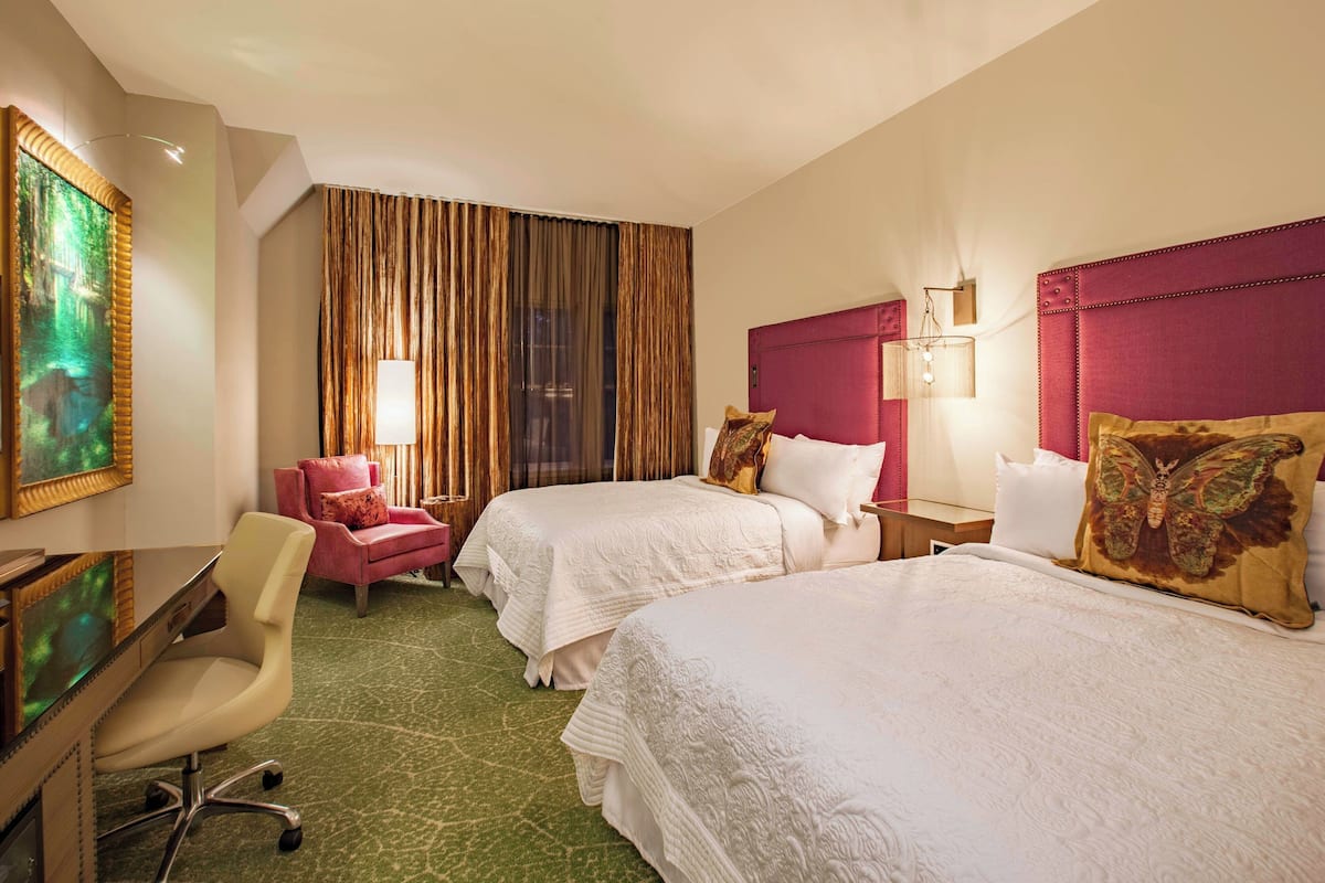 Une chambre d’hôtel avec deux lits, un bureau, une chaise, un tableau et une fenêtre avec des rideaux.