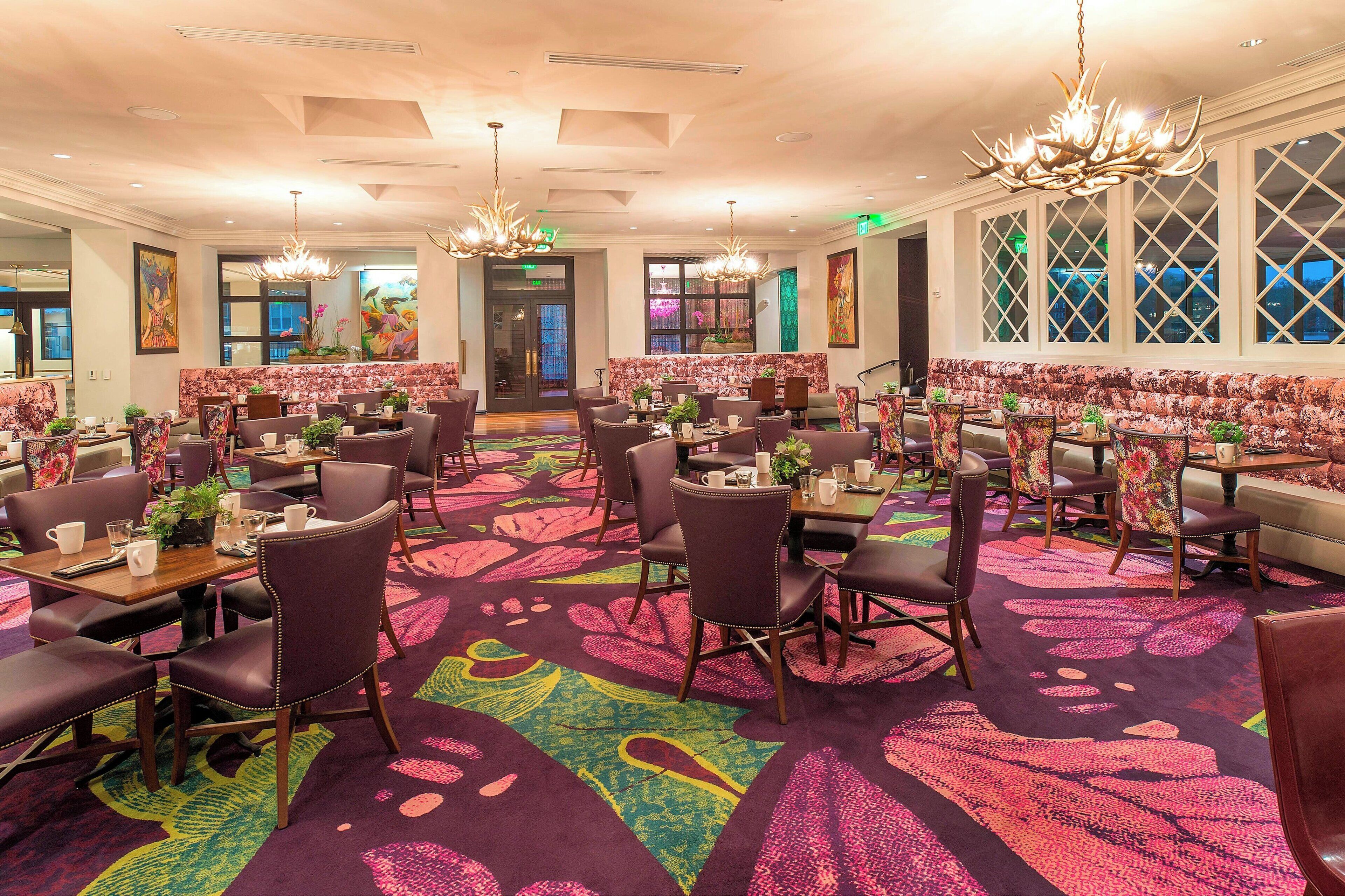 A hotel dining area with multiple tables and chairs, large windows, and a colorful carpet.