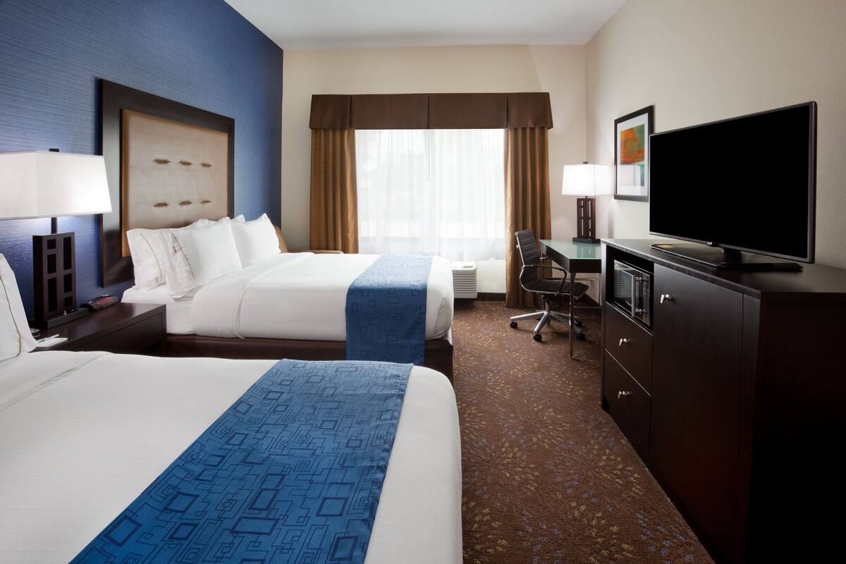 A hotel room with two beds, a desk, a television, and a large window with curtains.