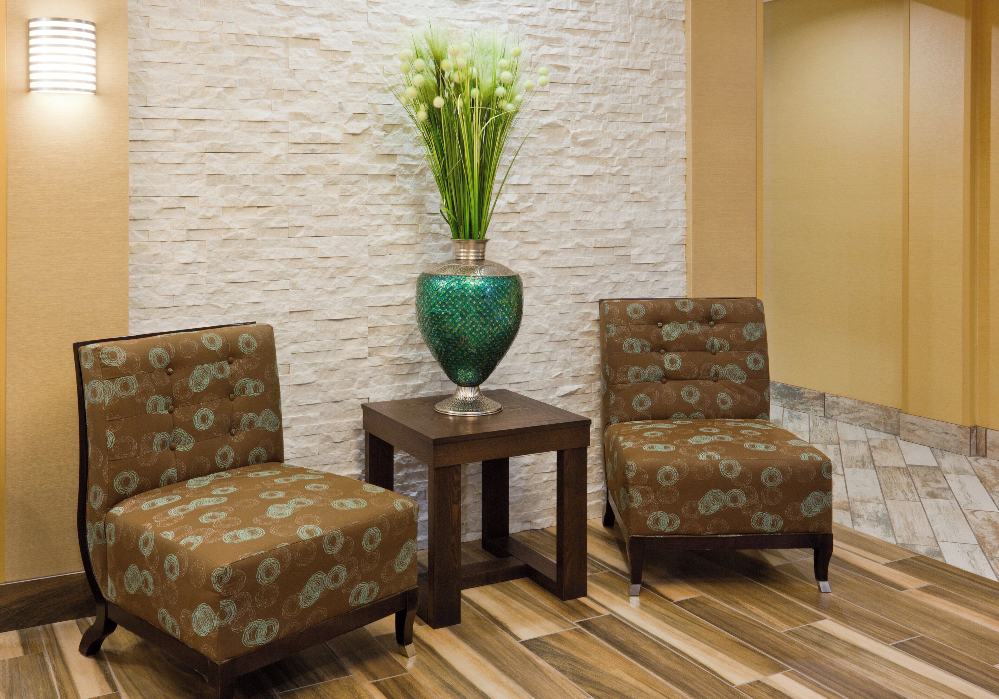 A seating area with two upholstered chairs and a small table, a decorative vase with green foliage, and a textured wall.