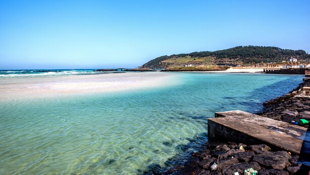 A beach with clear water, a rocky shoreline, and a distant hill with buildings.