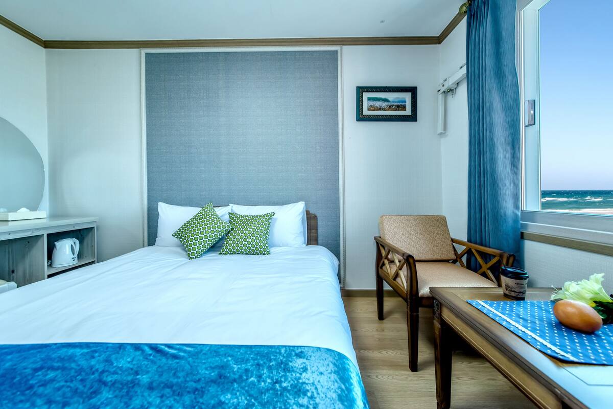 Une chambre d’hôtel avec un lit, une chaise, une table avec un set de table, un tableau au mur et une vue sur la mer par la fenêtre.