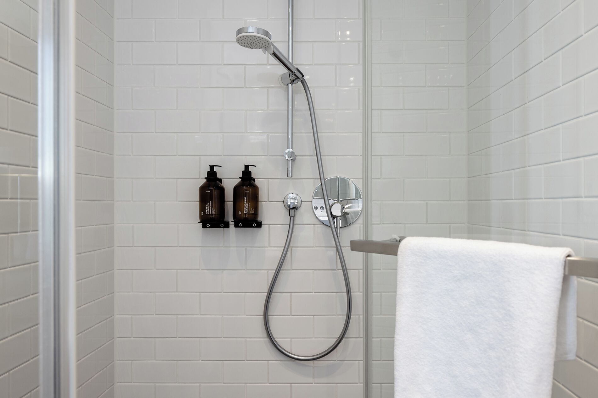 Eine Dusche mit Duschbrause und Wandarmatur. Auf einem Regal stehen zwei Flaschen Shampoo und Conditioner. Ein weißes Handtuch hängt an einem Ablagegestell.