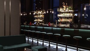 A bar area with a modern design, featuring a bar counter, stools, and a seating area with sofas and tables. - Hotel AMANO Grand Central (Berlin)