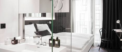 Una habitación de hotel moderna con una cama grande, cabina de ducha con paredes de vidrio, tocador blanco con artículos de aseo y vistas a la ciudad a través de amplias ventanas.