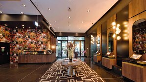 A lobby with a patterned carpet, a reception desk, a large vase with flowers, and a wall with a mural. - Hotel AMANO Grand Central (Berlin)