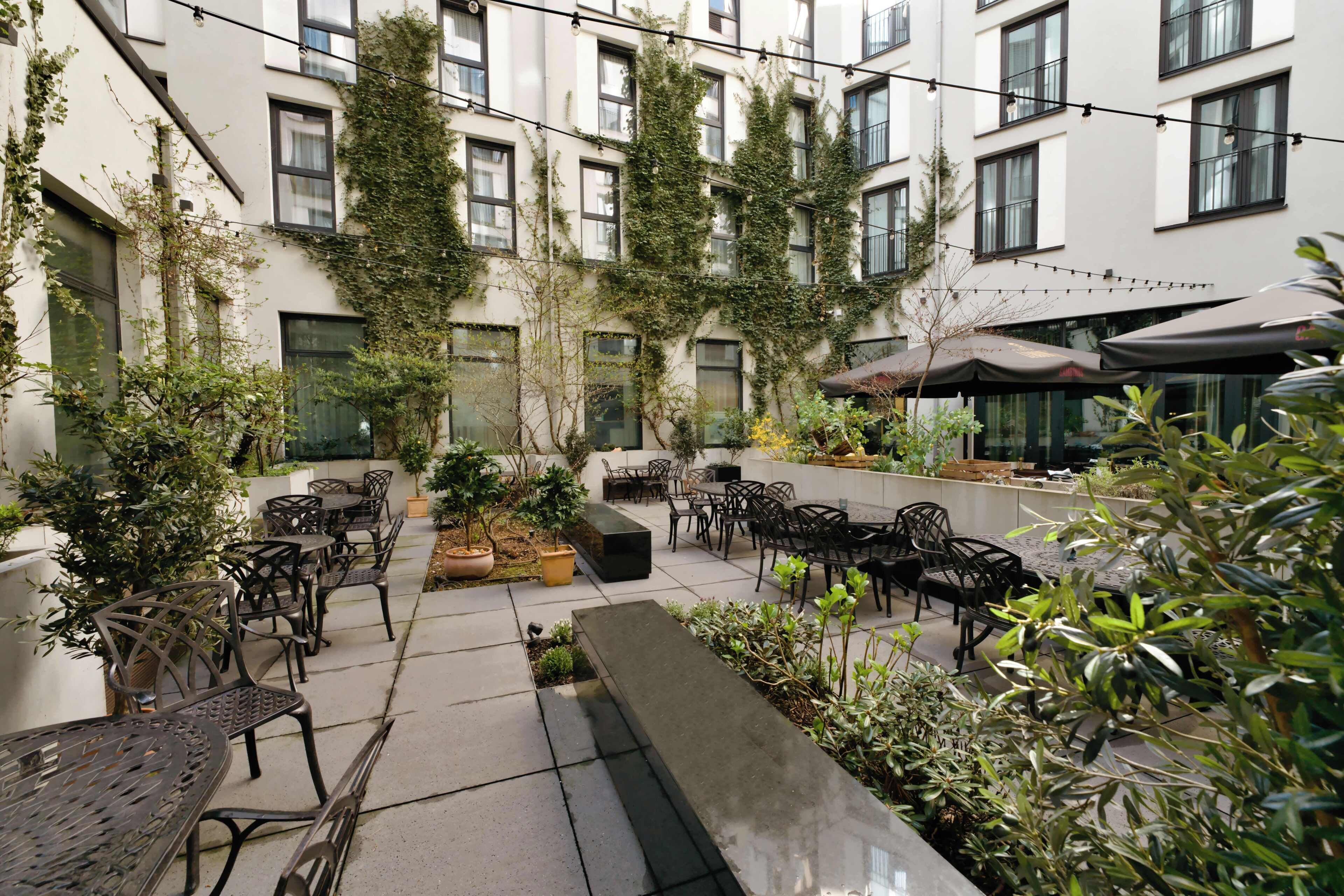 Un patio con mesas al aire libre, plantas en macetas y un edificio con varias ventanas.