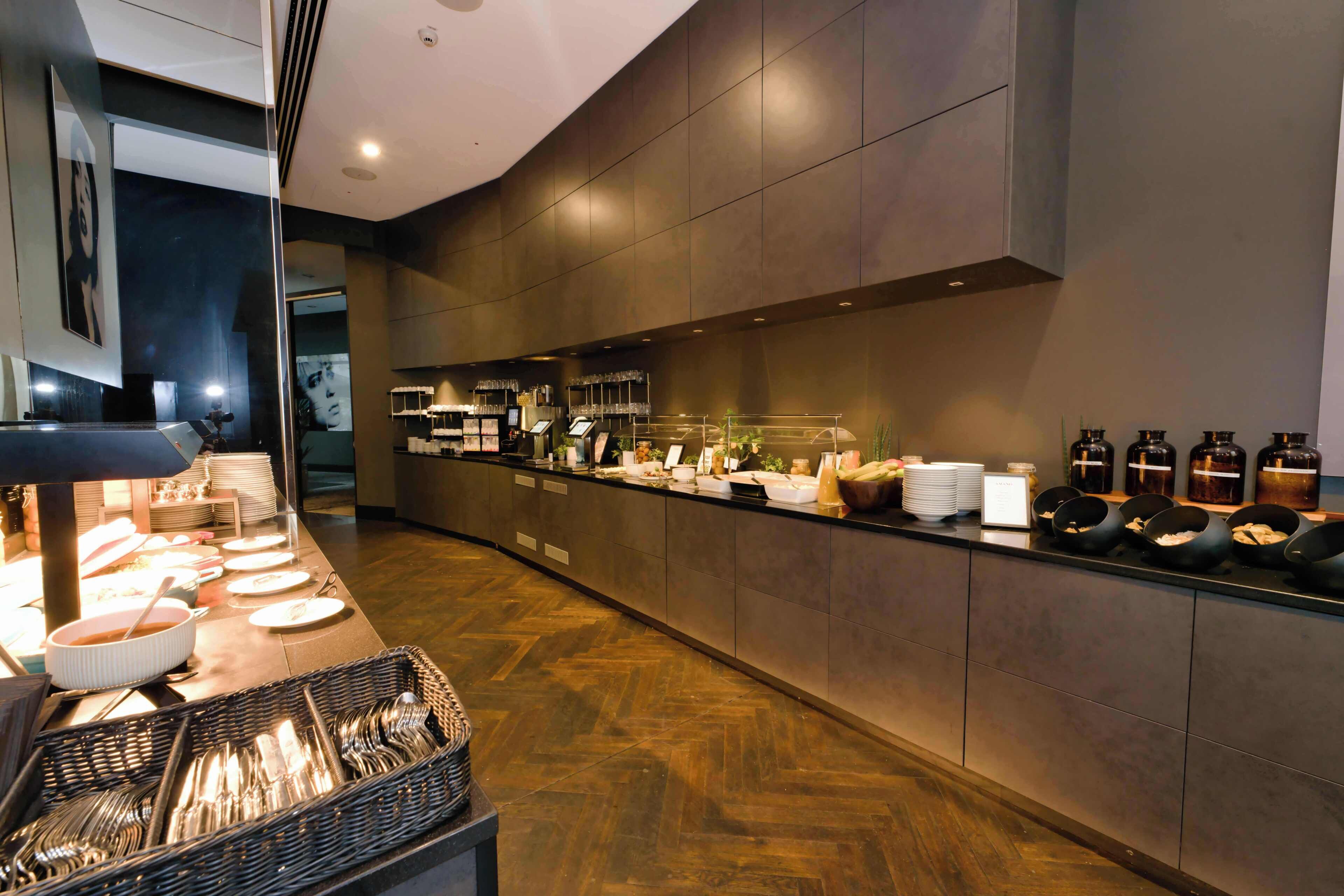 A modern hotel lobby with a long reception desk, a buffet area, and a large mirror reflecting the interior.