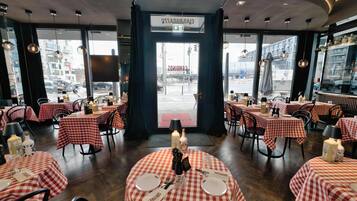 Un restaurant avec des tables rondes recouvertes de nappes à carreaux rouges et blancs, chacune agrémentée d’assiettes, de couverts et de bougies.