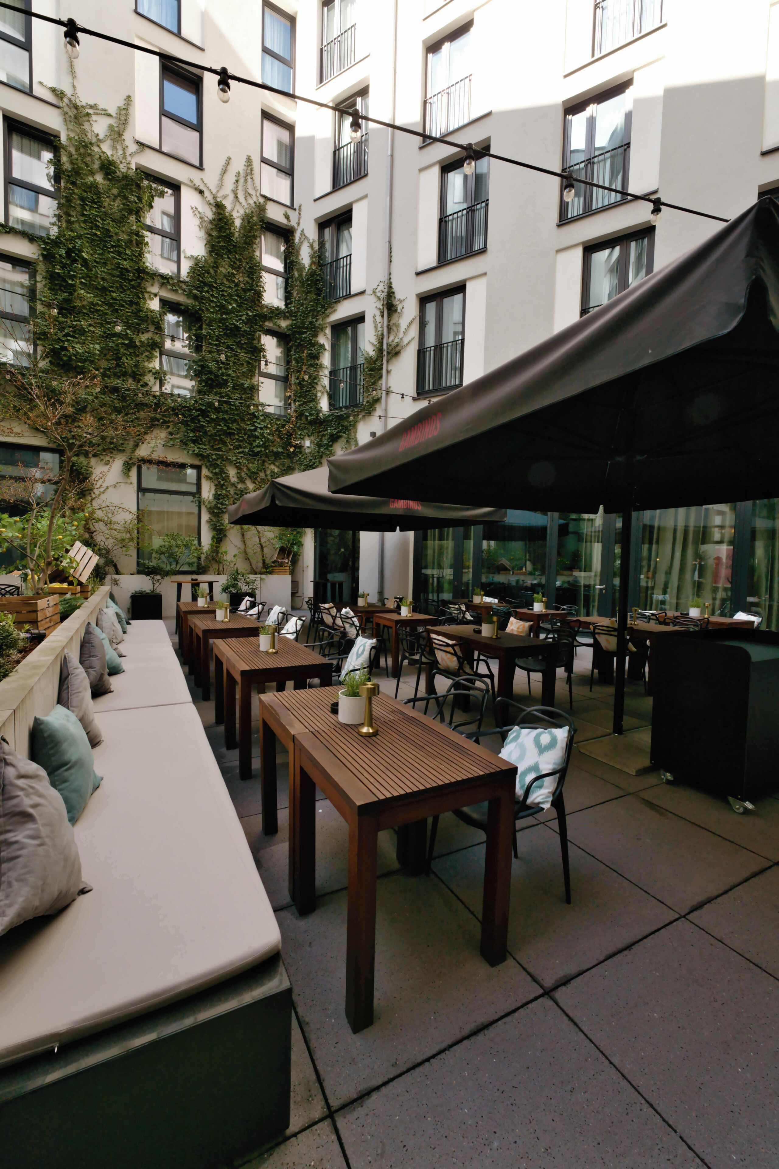 Een terras met houten tafels en banken, omringd door een gebouw met balkons en begroening.