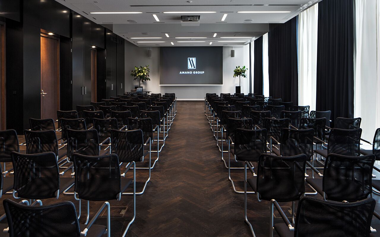 Ein Konferenzraum mit Reihen schwarzer Stühle, einer Projektionsleinwand mit dem Logo der Amano Group und zwei Blumentöpfen.