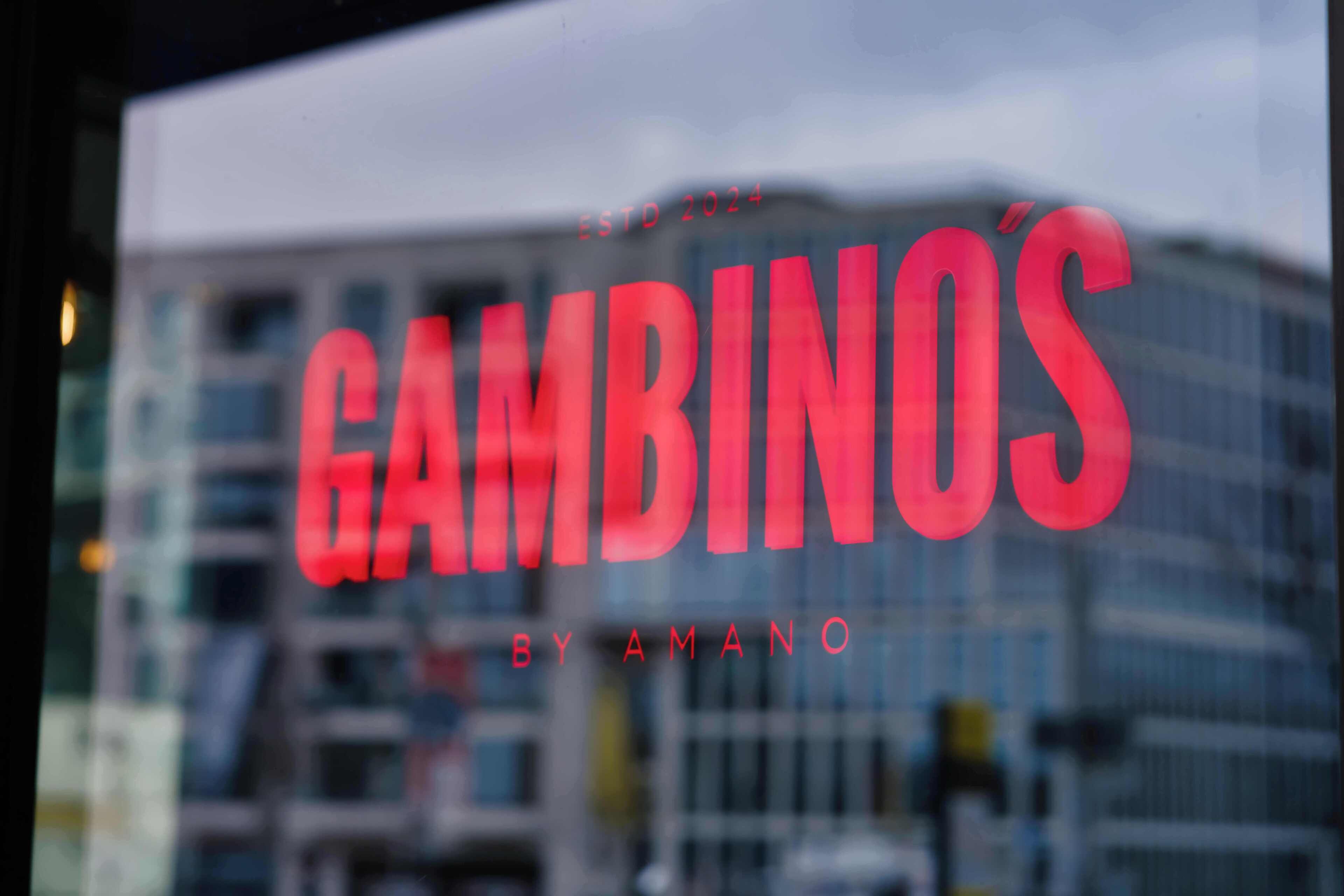Ikkunassa näkyy teksti ”GAMBINOS BY AMANO”, ja taustalla on epätarkka kuva rakennuksista.