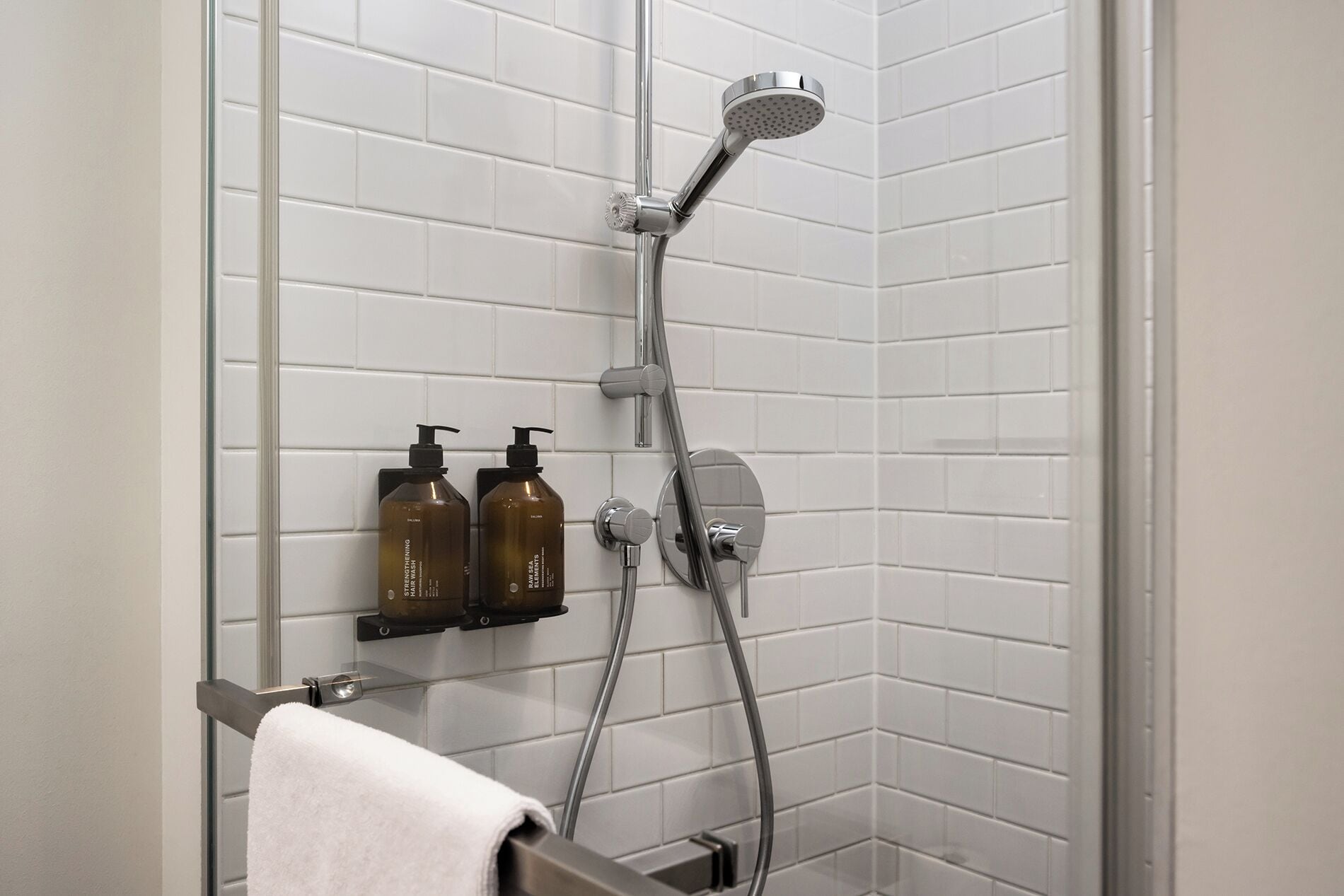 A shower with a handheld showerhead, a wall-mounted faucet, and two bottles of shampoo on a shelf.