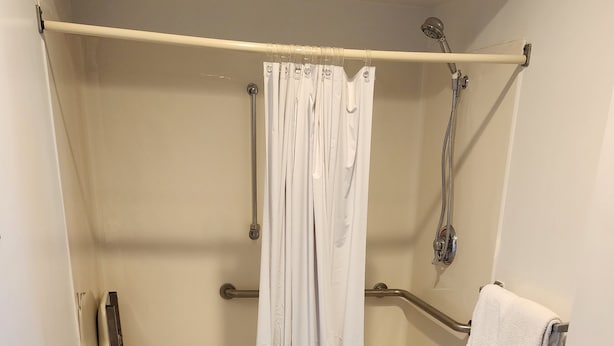 A shower area with a white curtain, grab bars, a towel rack, and a tiled floor. - Nittany Budget Motel (State College)