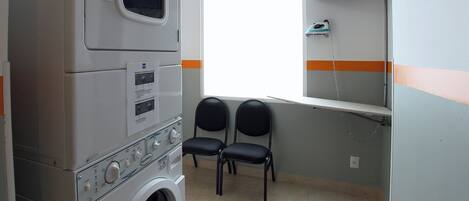 A laundry room with stacked washers and dryers, two chairs, a window, and a TV mounted on the wall.