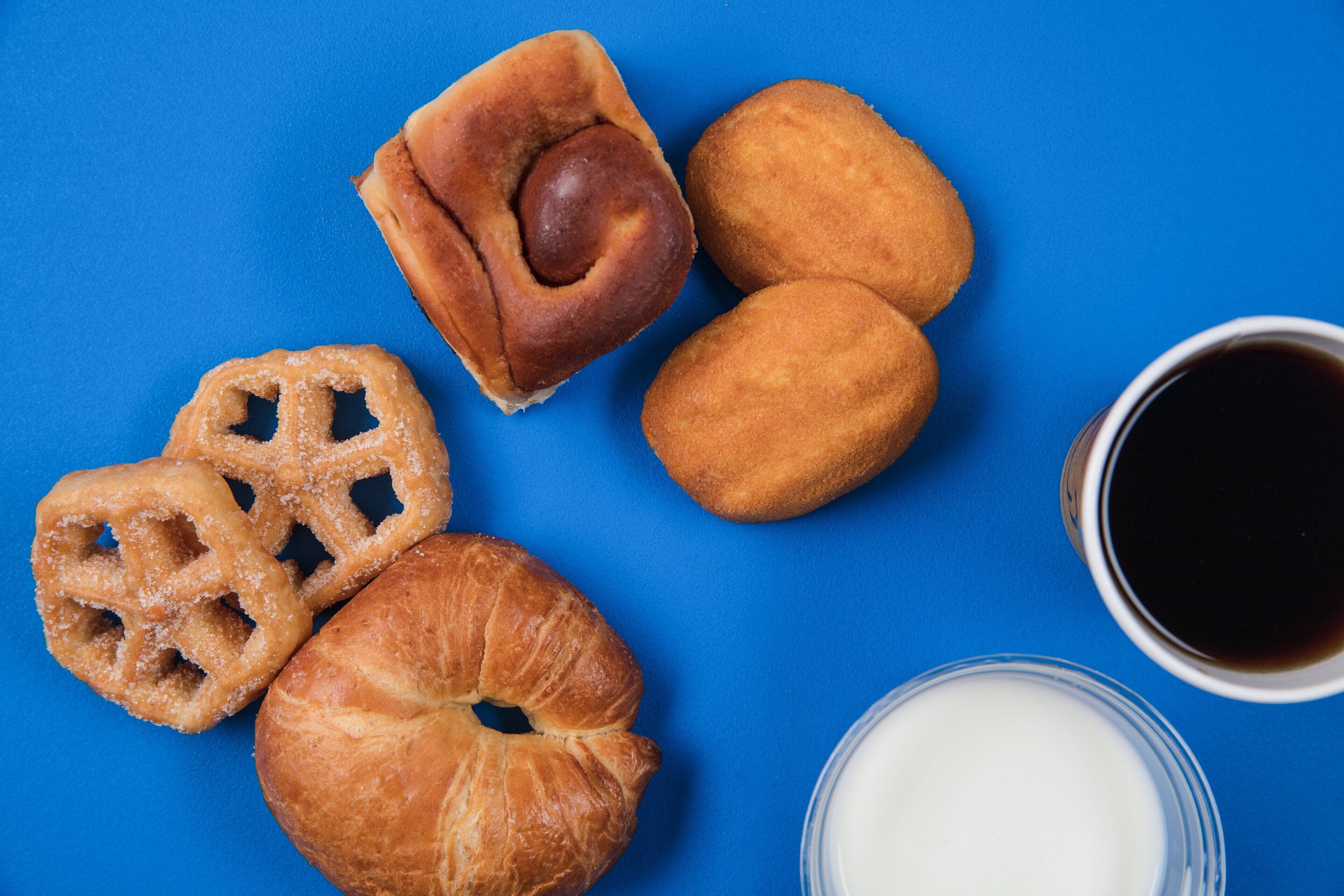 Diversos doces, incluindo um croissant, donut e bagel, acompanhados de uma xícara de café e um copo de leite sobre um fundo azul.