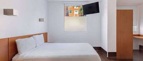 Une chambre d’hôtel moderne avec un grand lit, un téléviseur à écran plat fixé au mur, une petite fenêtre et une table de chevet en bois.