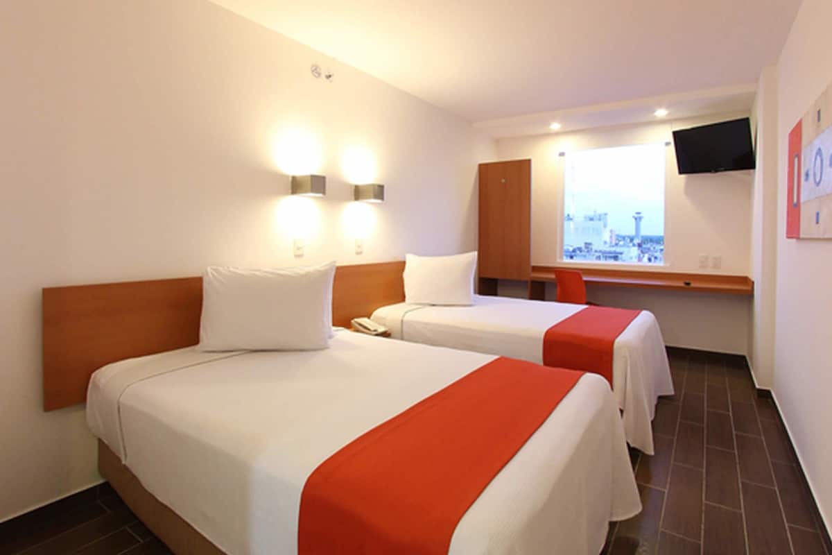 A hotel room with two beds, a TV mounted on the wall, and a window with a city view.