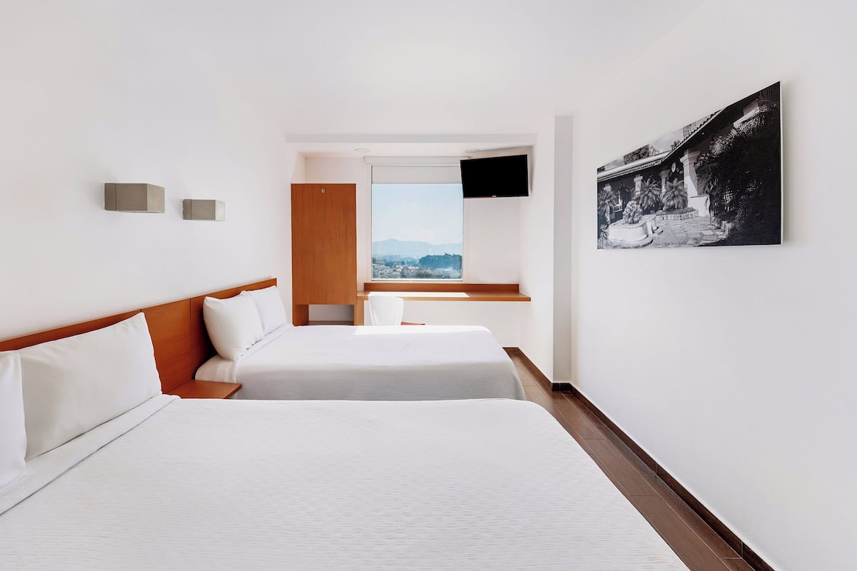 A hotel room with two beds, a wooden headboard, a flat-screen TV mounted on the wall, and a large window offering a cityscape view.