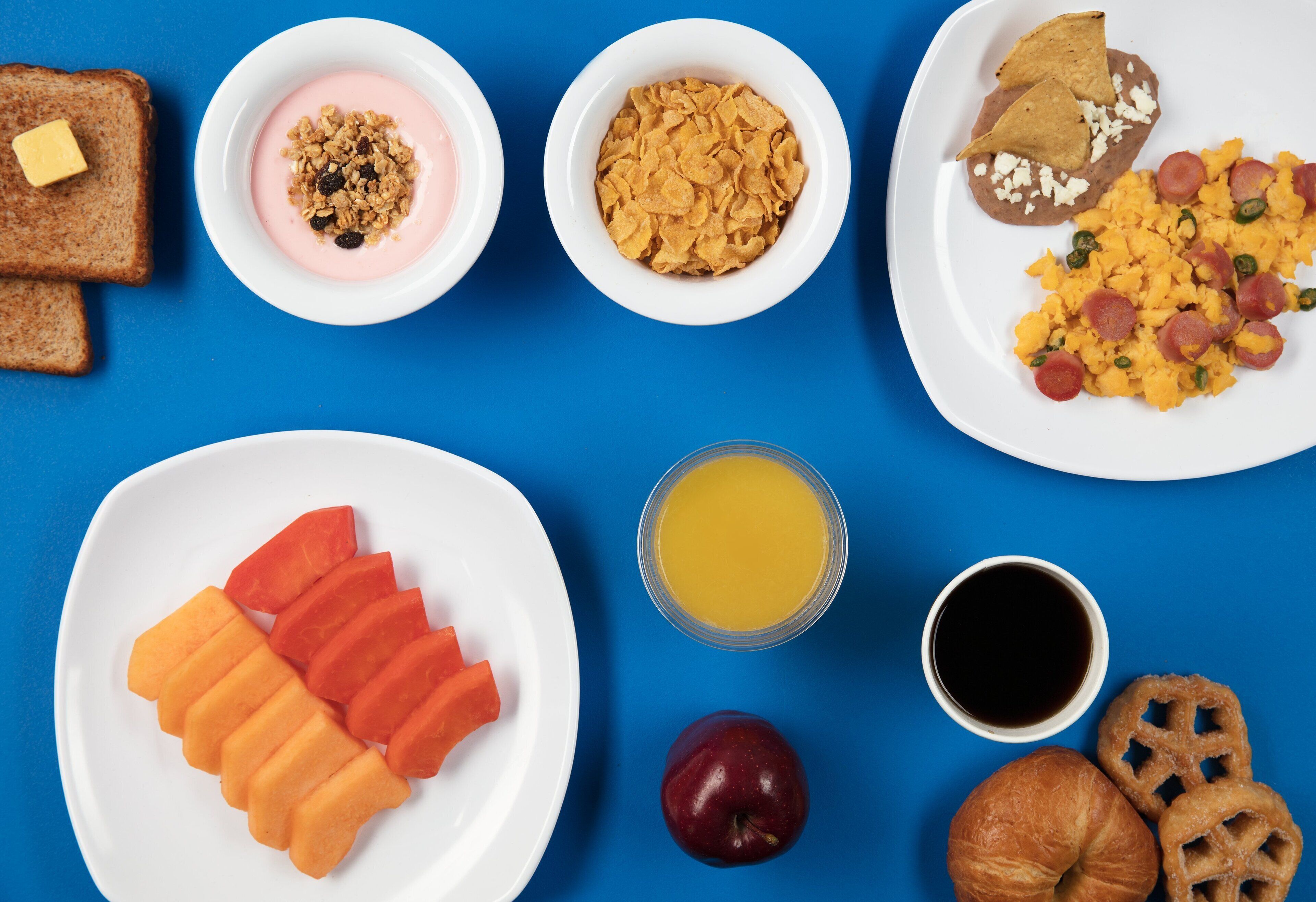 Una mesa de desayuno con tostadas, yogur, cereales, frijoles, huevos, salchicha y fruta en platos.