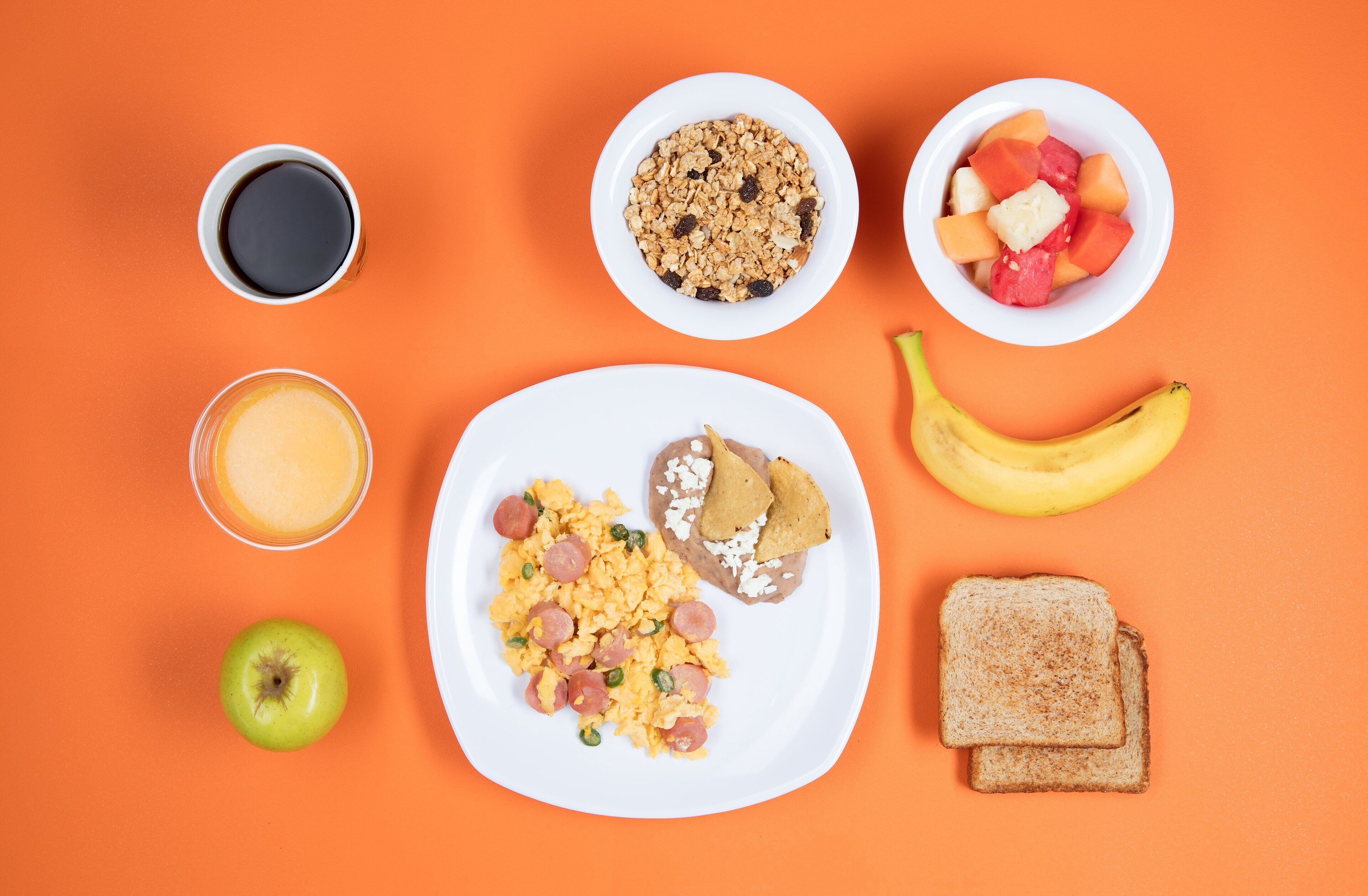 A breakfast spread on an orange table including scrambled eggs, toast, a banana, a cup of coffee, a bowl of cereal, a bowl of fruit, and a glass of orange juice.