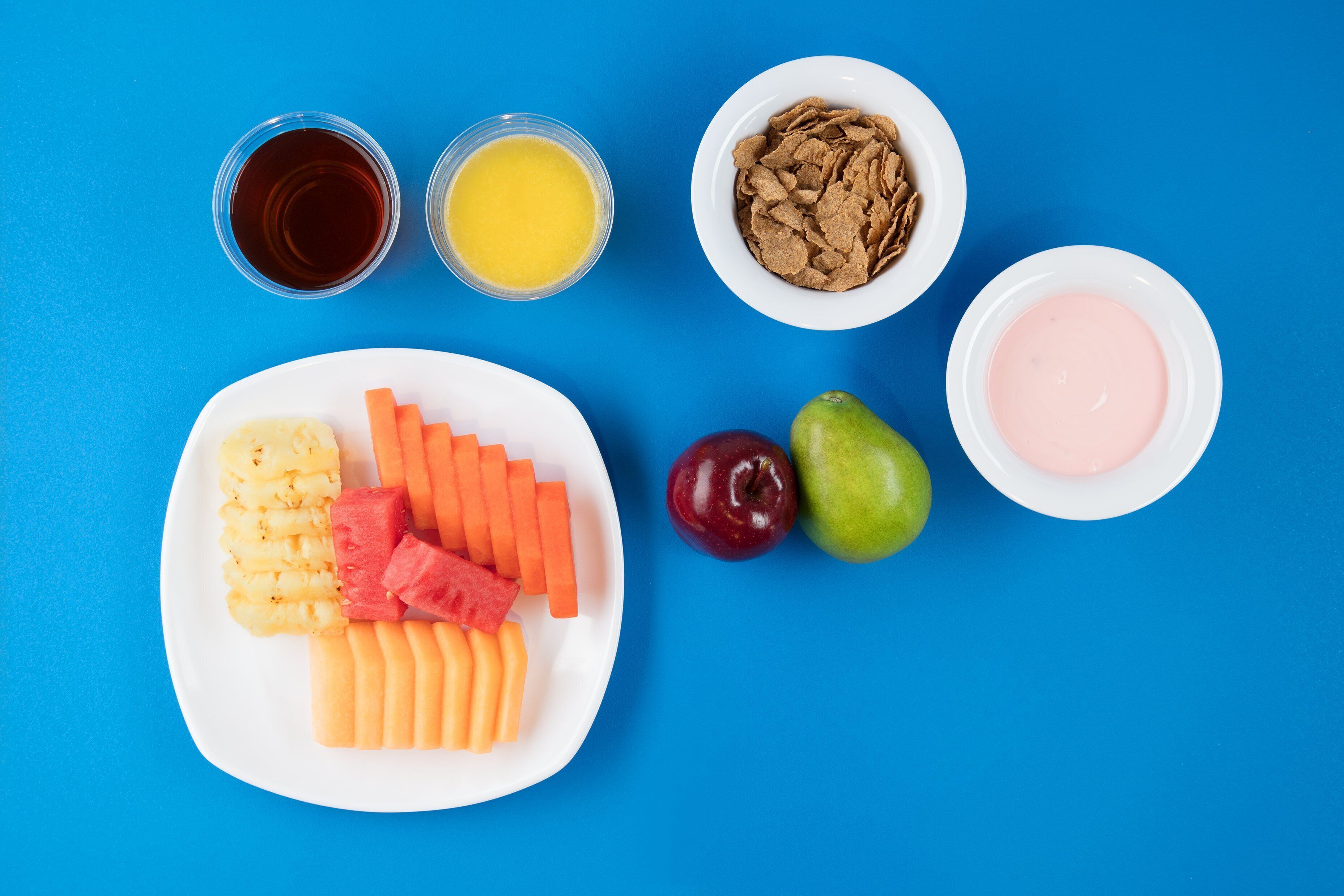 Un plato con frutas y verduras cortadas, dos vasos de jugo, un tazón de cereal y un tazón de yogur sobre un fondo azul.