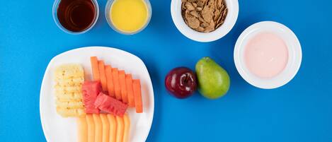 Un plato con frutas y verduras cortadas, dos vasos de jugo, un tazón de cereal y un tazón de yogur sobre un fondo azul.