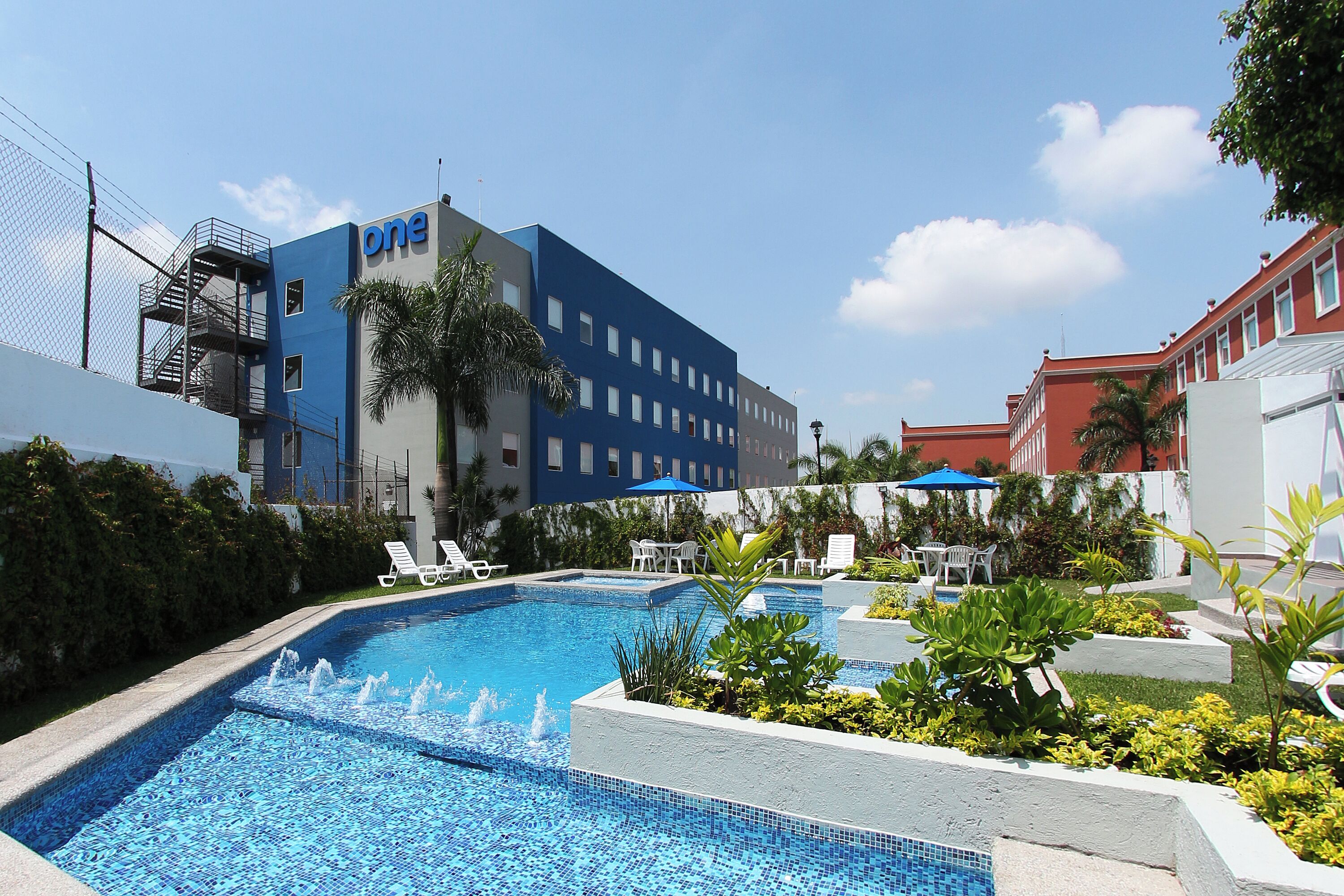 A hotel pool area with a blue building labeled 'one' in the background, surrounded by greenery and lounge chairs.