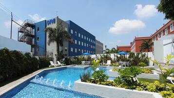 A hotel pool area with a blue building labeled 'one' in the background, surrounded by greenery and lounge chairs.