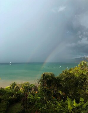 Un paysage côtier avec un arc-en-ciel, une végétation luxuriante et une mer calme où flottent des bateaux.