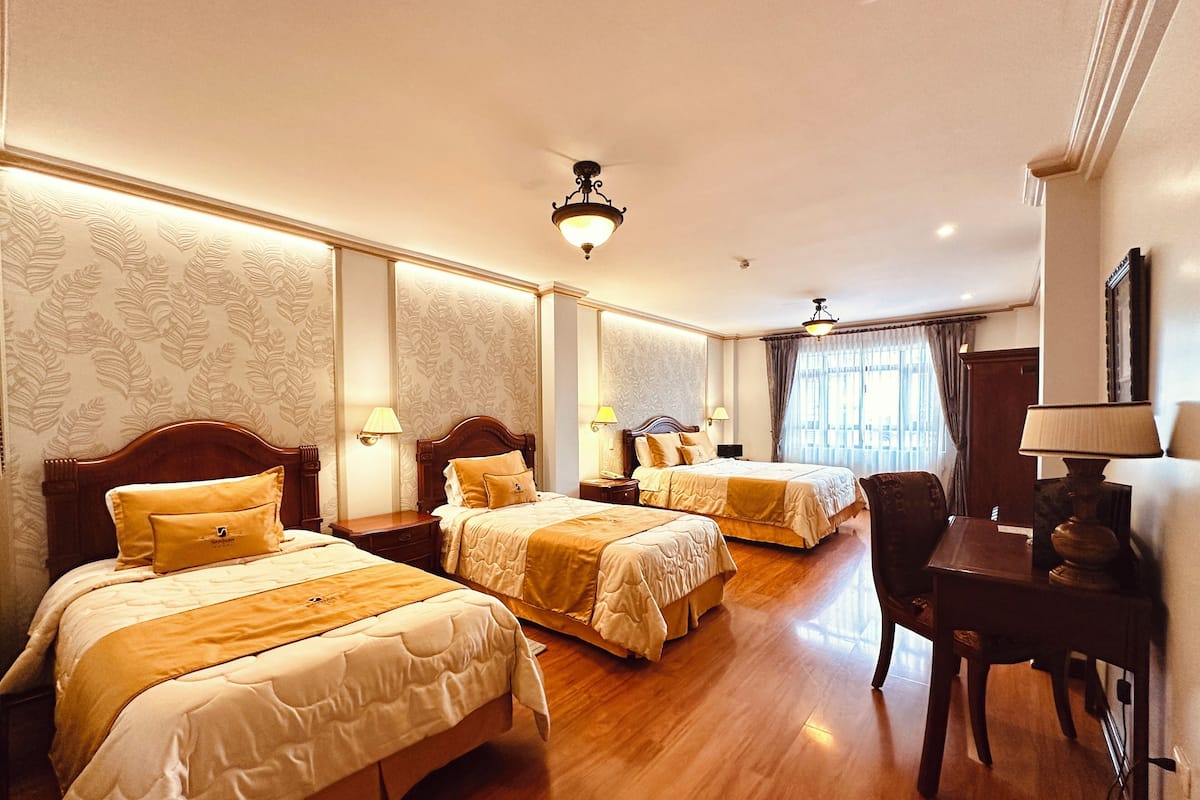 A hotel room with three double beds, wooden flooring, and a desk with a chair.