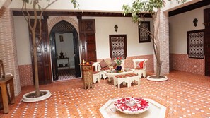 Un patio con piso de baldosas, una fuente con pétalos de rosa, una zona de estar con cojines y una puerta de madera con diseños intrincados.