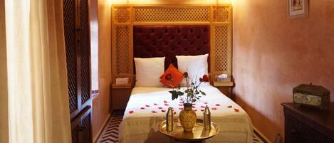 A hotel room with a red upholstered headboard, white bedding, a small bedside table with a vase of flowers, and a decorative tray.