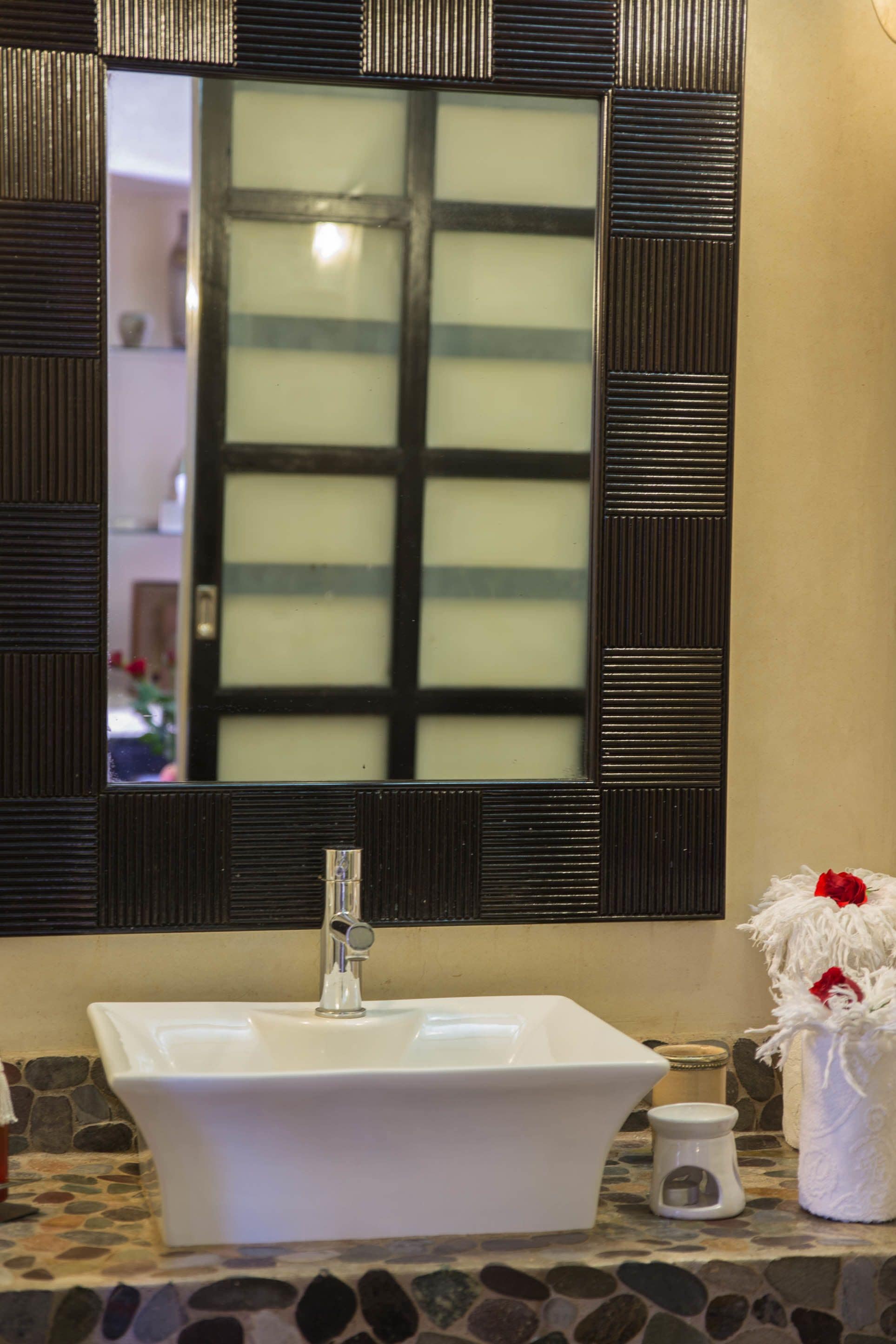Un baño con un lavamanos blanco, un espejo con marco decorativo y un pequeño jarrón con una flor roja.