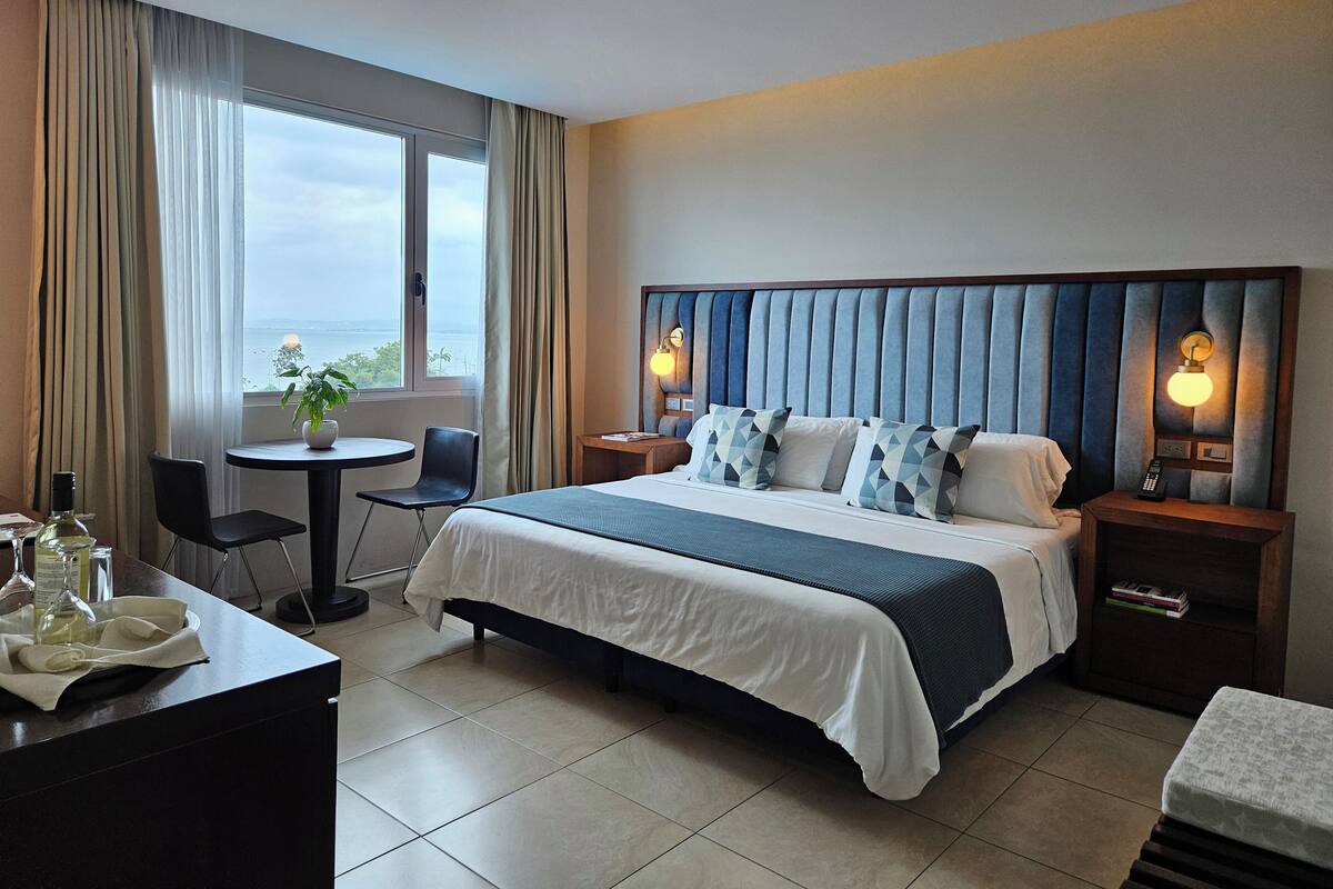 A hotel room with a large bed, a bedside table with a lamp, a small round table with a chair, a desk with a chair, and a view of the sea through the window.
