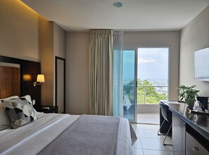 A hotel room with a bed, a desk, a chair, a lamp, a plant, and a view of the sea and cityscape through the window. - Hotel Perla Spondylus (Manta)
