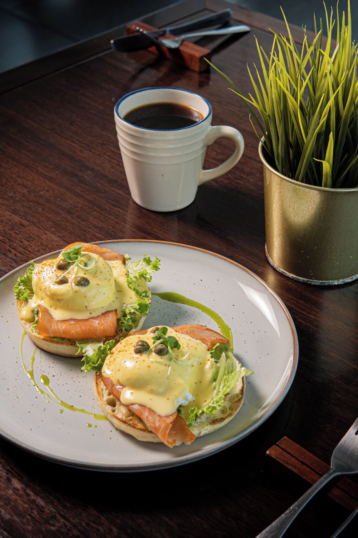 A plate of eggs benedict with hollandaise sauce and capers, a white coffee cup, and a small potted plant on a wooden table.