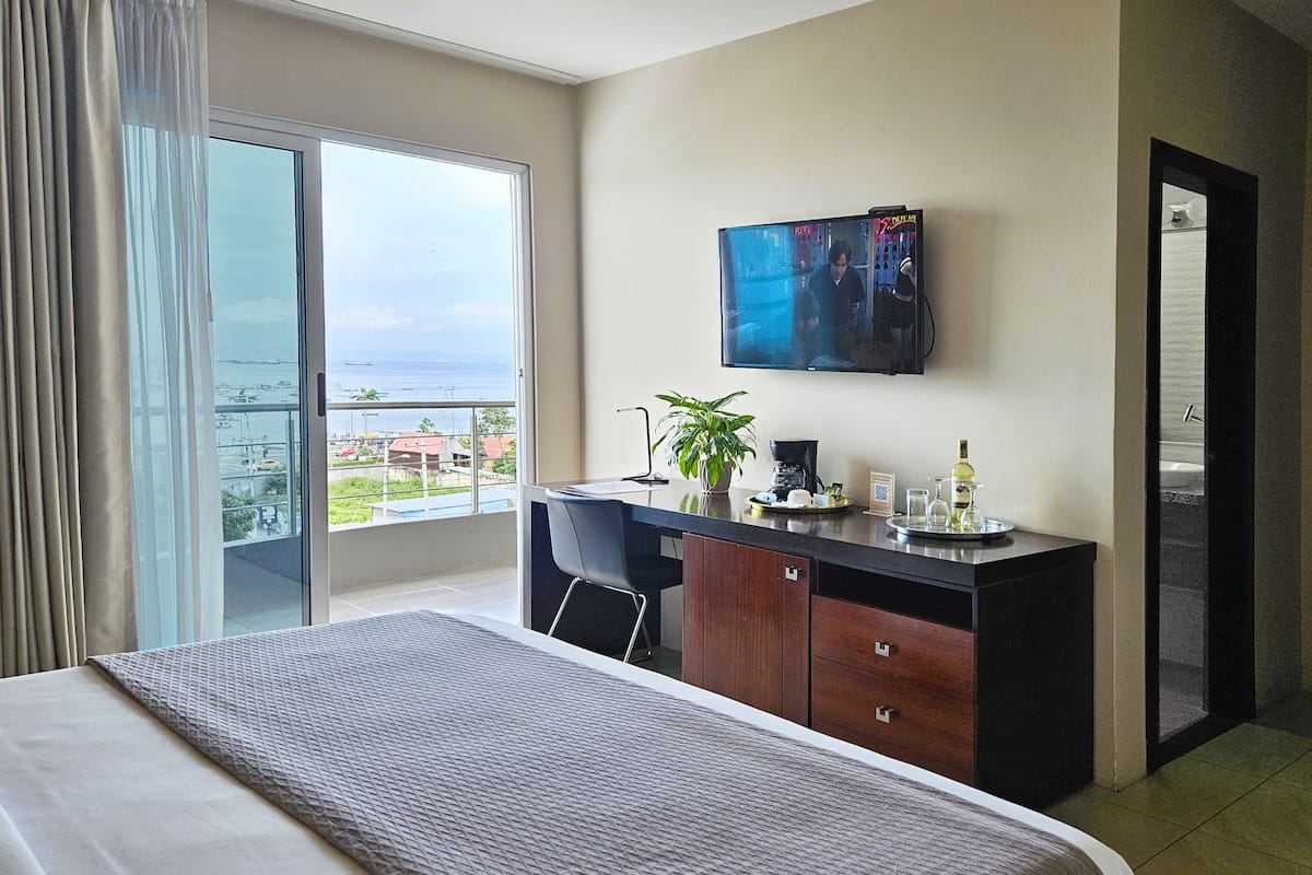 A hotel room with a bed, a desk with a plant, a TV, and a view of the ocean.