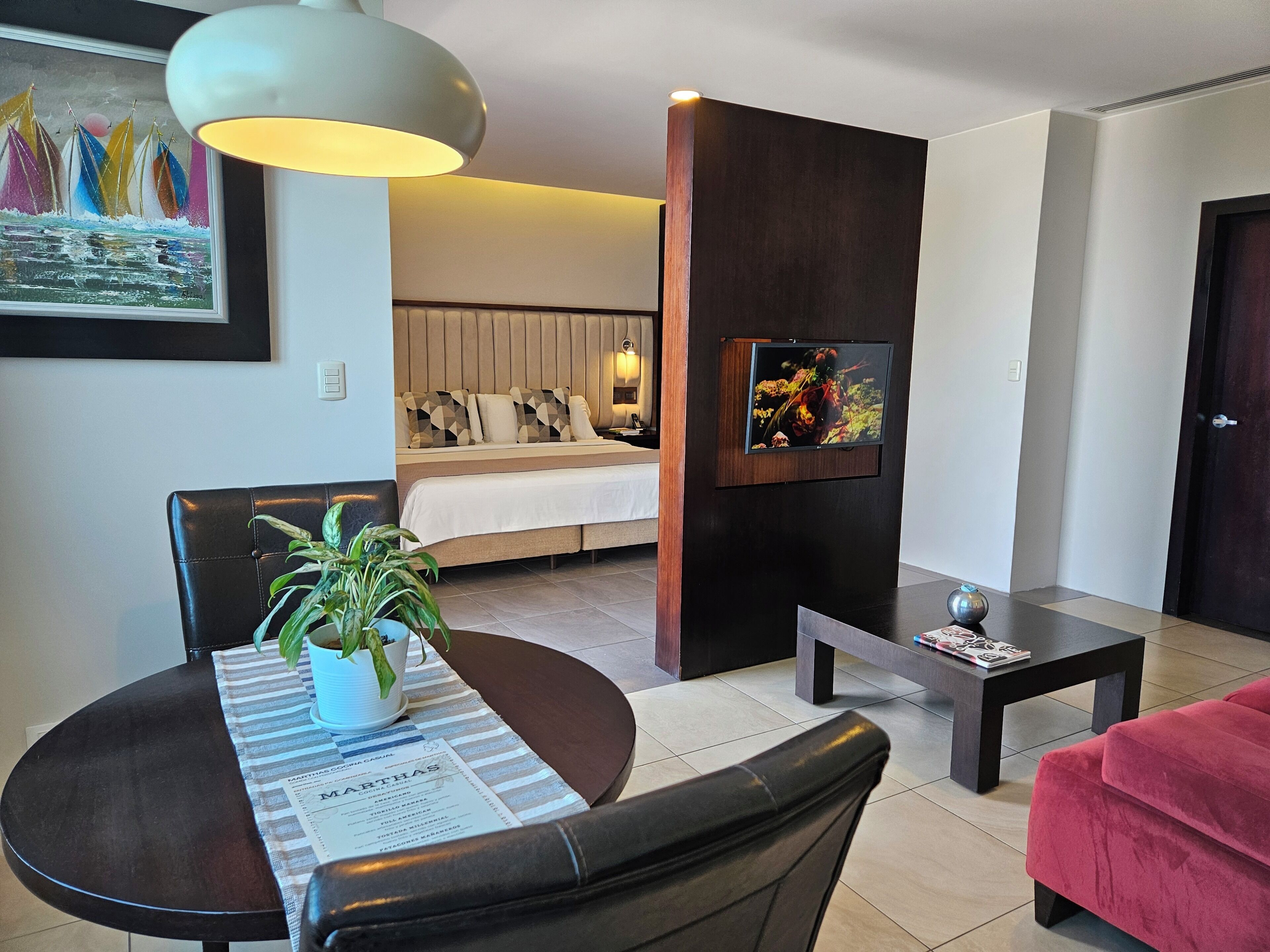 A modern living room with a dining area, a sofa, a TV mounted on a wooden panel, and a potted plant on the dining table.