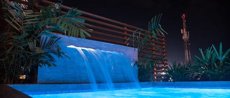 A nighttime pool area with a waterfall feature, surrounded by tropical plants and a wooden structure.